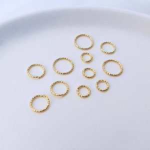 May include: Assortment of 10 gold-toned jump rings in various sizes. The rings are all round and have a textured, braided design.