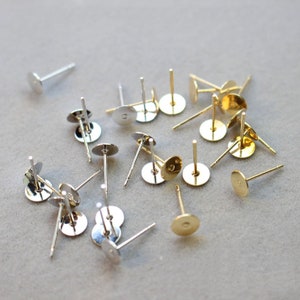 Op de afbeelding: Een verzameling gouden en zilveren oorbelstiften met platte ronde achterkanten. De stiften zijn verspreid over een lichtgrijze ondergrond.