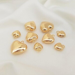 Puede incluir: Una colección de dijes en forma de corazón de diferentes tamaños, de color dorado. Los dijes tienen un acabado liso y brillante y presentan un pequeño bucle en la parte superior para sujetarlos a joyas u otras manualidades.