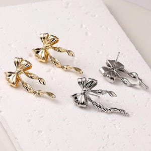 May include: A pair of gold and silver bow earrings with twisted ribbon details. The earrings are designed with a bow shape and have a post back closure.