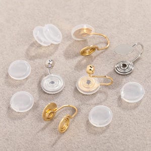 May include: A collection of gold and silver-coloured clip-on earring components and clear, round earring backs are arranged on a neutral surface. The clip-on components have a coiled design and a curved arm for attachment. The earring backs are made of clear, soft plastic.