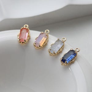 May include: Four gold-tone charms with different colored rectangular glass stones. The charms are on a white surface.