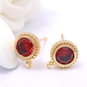 Puede incluir: Un par de pendientes de botón dorados. Cada pendiente presenta una piedra preciosa redonda de color rojo intenso engastada en un borde texturizado en forma de cuerda. Los pendientes tienen un poste y un pequeño lazo.