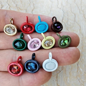 May include: A collection of eleven small oval charms featuring faceted crystals set in colorful metal frames. The charms include various colors such as red, blue, green, pink, yellow, white, and gold, each with a small loop at the top for attachment.