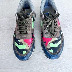 May include: A pair of camouflage sneakers with a mix of pink, green, and brown patterns. The sneakers have dark green laces, a blue sole, and a gray padded tongue. The shoes are designed for casual wear.