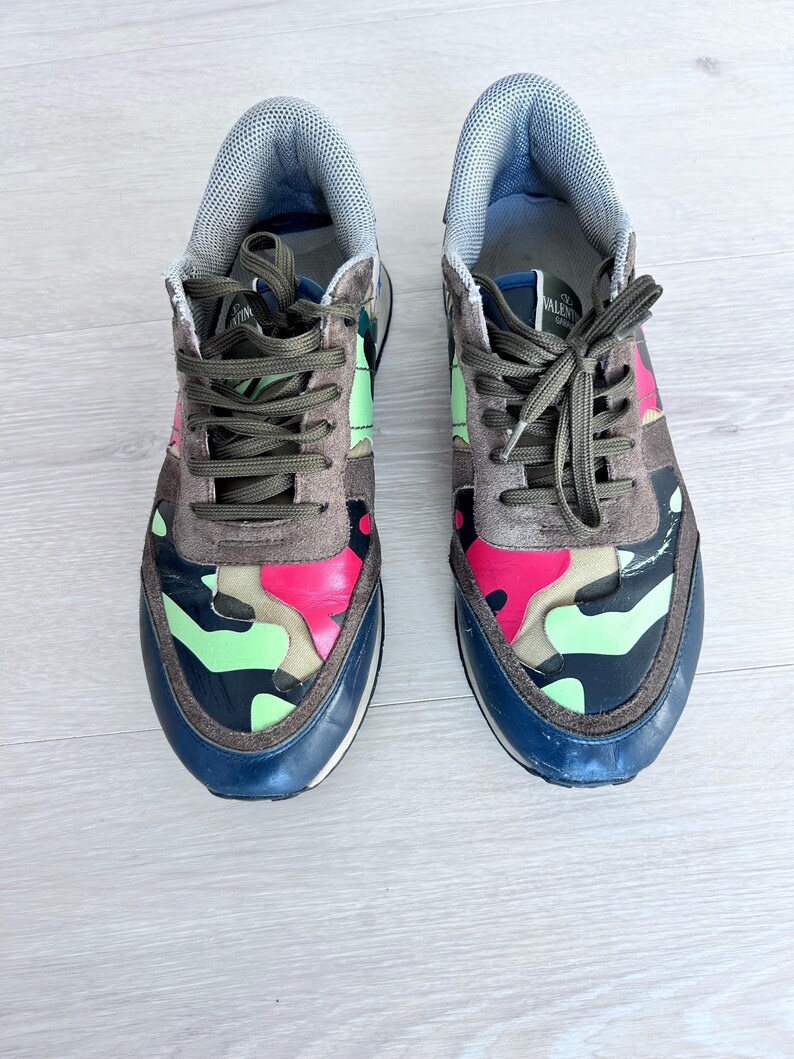 May include: A pair of camouflage sneakers with a mix of pink, green, and brown patterns. The sneakers have dark green laces, a blue sole, and a gray padded tongue. The shoes are designed for casual wear.