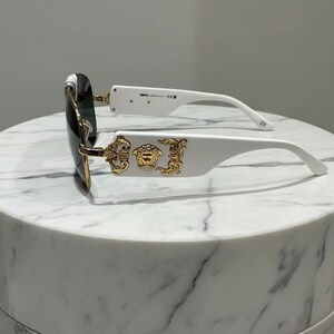 Puede incluir: Gafas de sol blancas con detalles dorados y lentes oscuras. Las monturas presentan un emblema dorado en las patillas. Las gafas de sol est&aacute;n sobre una superficie de m&aacute;rmol blanco.