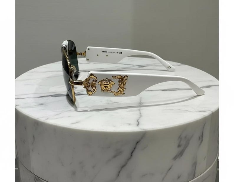 Puede incluir: Gafas de sol blancas con detalles dorados y lentes oscuras. Las monturas presentan un emblema dorado en las patillas. Las gafas de sol est&aacute;n sobre una superficie de m&aacute;rmol blanco.
