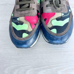 May include: Pair of colorful sneakers with a camouflage design. The shoes feature a combination of pink, green, and black accents on the toe and side panels, with a navy blue toe cap and gray suede detailing. The laces are olive green.