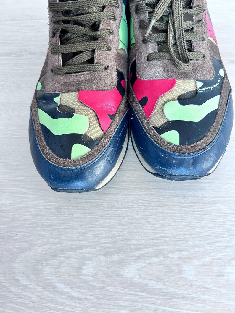 May include: Pair of colorful sneakers with a camouflage design. The shoes feature a combination of pink, green, and black accents on the toe and side panels, with a navy blue toe cap and gray suede detailing. The laces are olive green.