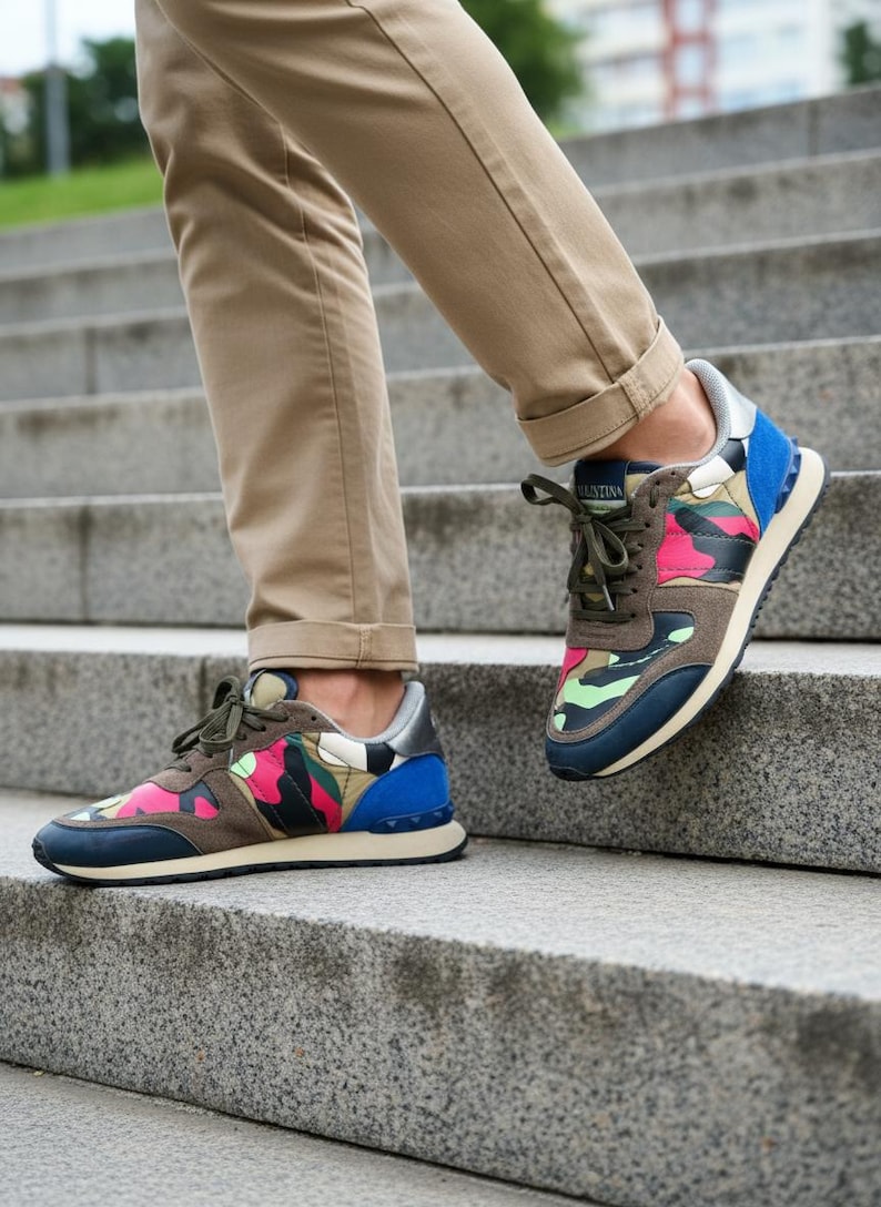 May include: Multi-colored camouflage sneakers with blue, pink, green, and brown accents. The sneakers have a navy blue sole and a silver heel. The laces are olive green. The sneakers are on a person walking down a set of concrete stairs.