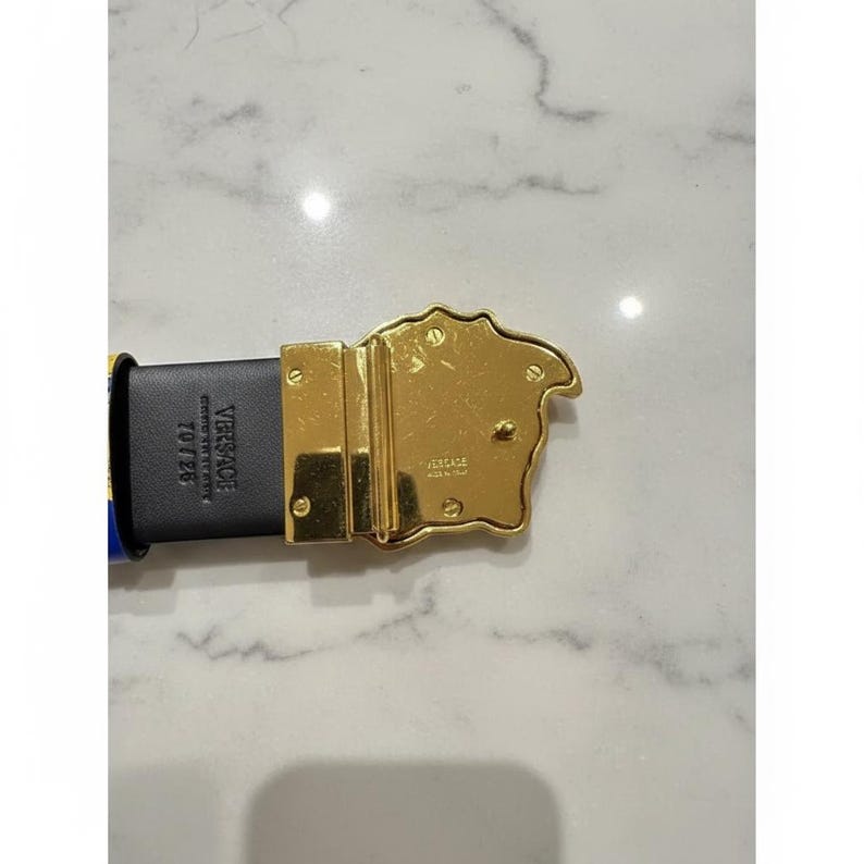 May include: A black leather belt with a gold-coloured buckle. The buckle features a Medusa head design and the brand name "Versace". The belt has blue and yellow accents.