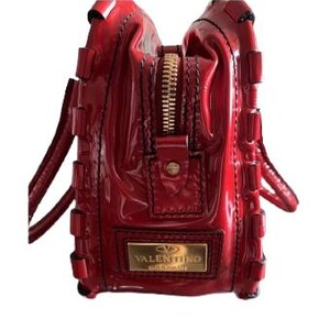 May include: A glossy red handbag with a gold zipper and handles. The bag features a rectangular gold plaque with the brand name "VALENTINO GARAVANI". The bag has a structured design with decorative stitching.