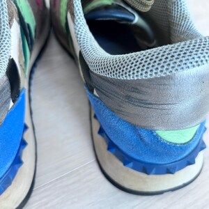 May include: Close-up of a pair of sneakers with a multi-colored design. The shoes feature a blue, gray, and white color scheme with a studded blue sole. The laces are olive green, and the interior is blue.