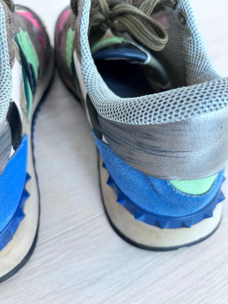 May include: Close-up of a pair of sneakers with a multi-colored design. The shoes feature a blue, gray, and white color scheme with a studded blue sole. The laces are olive green, and the interior is blue.