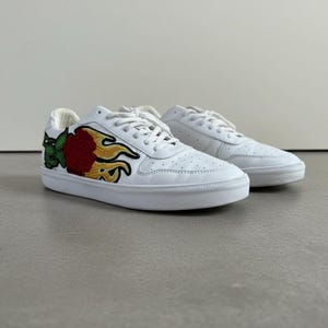 May include: A pair of white low-top trainers featuring a prominent embroidered patch on the side. The patch depicts a red rose surrounded by yellow and orange flames with green leaves, set against the clean white leather of the shoe upper.