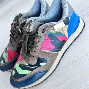 May include: A pair of colorful sneakers with a camouflage pattern. The shoes feature a mix of colors, including pink, green, blue, and brown. They have brown laces and a blue sole. The sneakers are made of suede and leather.