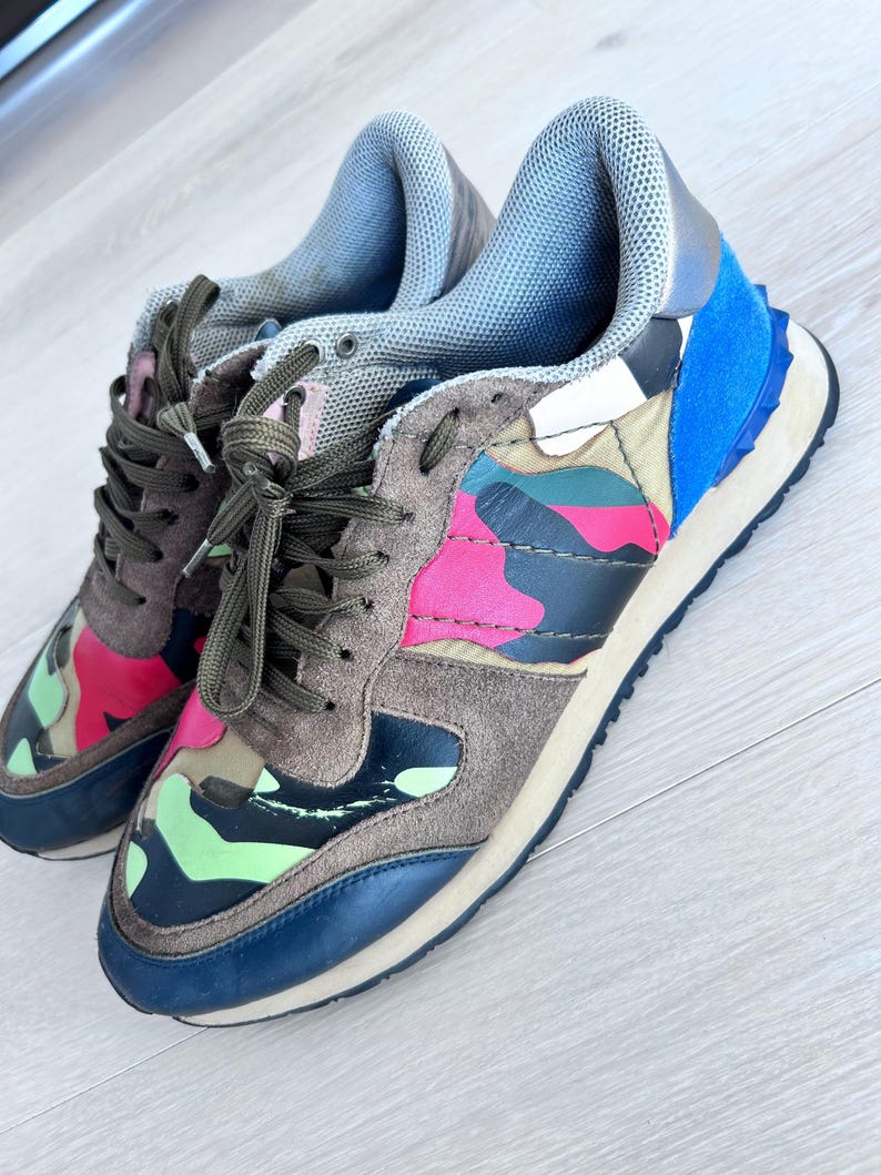 May include: A pair of colorful sneakers with a camouflage pattern. The shoes feature a mix of colors, including pink, green, blue, and brown. They have brown laces and a blue sole. The sneakers are made of suede and leather.