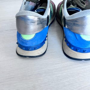 May include: A pair of colorful sneakers with a mix of blue, silver, and floral print accents. The shoes feature a blue heel counter, a light green detail, and a studded sole. The laces are dark green.