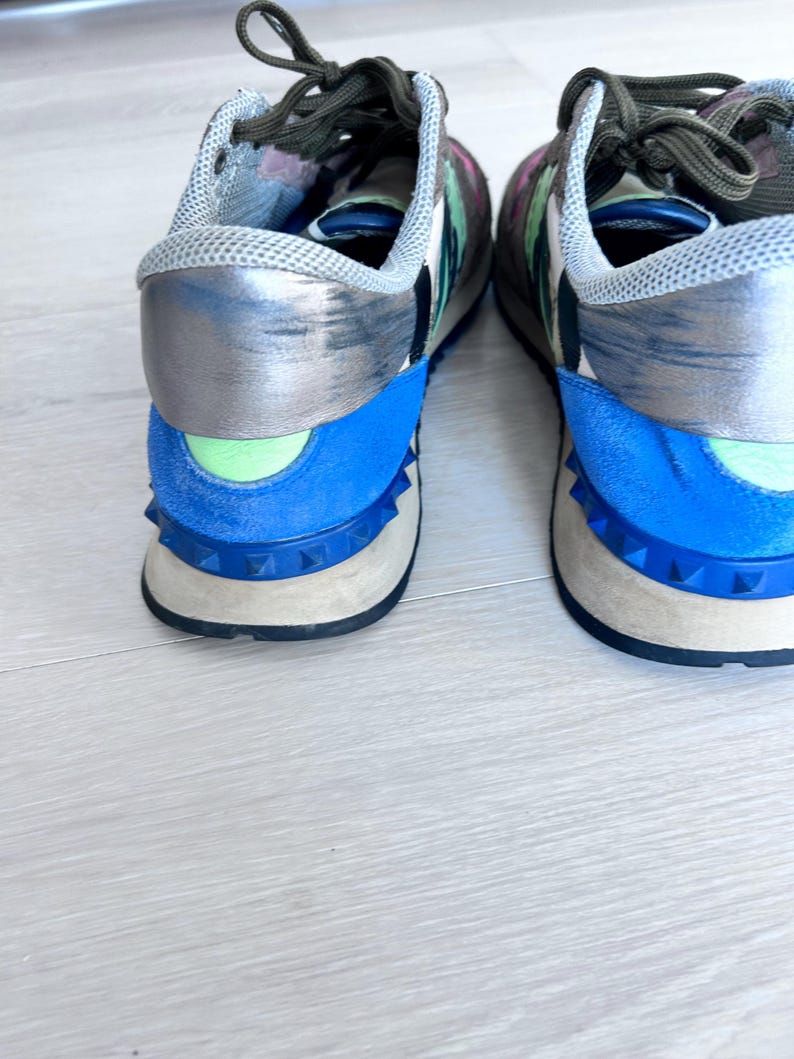 May include: A pair of colorful sneakers with a mix of blue, silver, and floral print accents. The shoes feature a blue heel counter, a light green detail, and a studded sole. The laces are dark green.