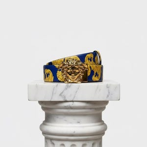 May include: A blue and gold patterned belt with a gold Medusa head buckle. The belt is displayed on a white marble pedestal. The belt's pattern features a repeating gold design against a blue background. The belt is a stylish accessory.