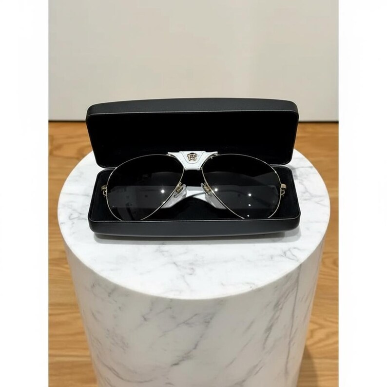 Puede incluir: Gafas de sol de aviador negras con montura dorada en un estuche negro abierto. Las gafas de sol tienen un puente plateado y un emblema decorativo. El estuche se encuentra sobre un soporte cil&iacute;ndrico blanco con vetas de m&aacute;rmol.