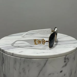 Puede incluir: Gafas de sol blancas con lentes negras. Las patillas presentan adornos dorados. Las gafas de sol est&aacute;n sobre una superficie de m&aacute;rmol blanco y gris.