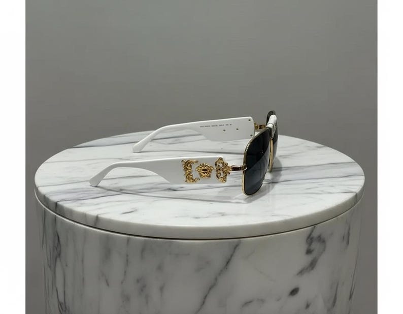 Puede incluir: Gafas de sol blancas con lentes negras. Las patillas presentan adornos dorados. Las gafas de sol est&aacute;n sobre una superficie de m&aacute;rmol blanco y gris.