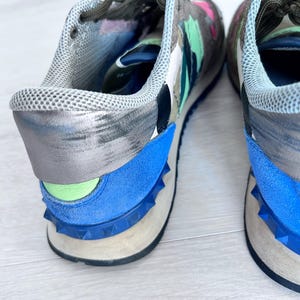 May include: A pair of colorful sneakers with a camouflage-like pattern in shades of gray, green, pink, and blue. The shoes have blue suede accents and a studded sole. The laces are dark green.