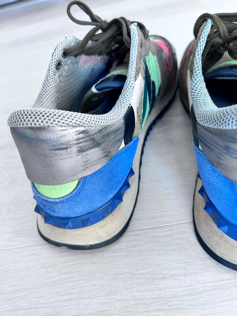 May include: A pair of colorful sneakers with a camouflage-like pattern in shades of gray, green, pink, and blue. The shoes have blue suede accents and a studded sole. The laces are dark green.