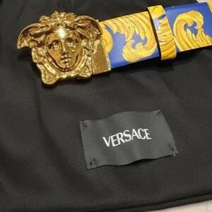 May include: A gold-coloured belt buckle featuring a Medusa head design, combined with a blue and gold patterned belt. The belt and buckle are on black fabric, with a tag that reads "VERSACE".