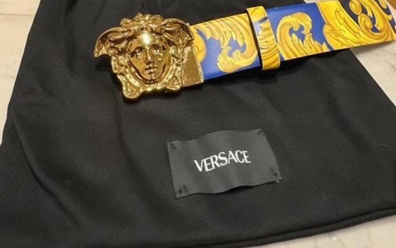 May include: A gold-coloured belt buckle featuring a Medusa head design, combined with a blue and gold patterned belt. The belt and buckle are on black fabric, with a tag that reads "VERSACE".