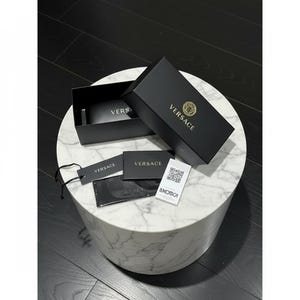 Puede incluir: Una caja negra de Versace con letras y logotipo dorados, abierta para revelar gafas de sol. Tambi&eacute;n hay etiquetas adicionales y un pa&ntilde;o de limpieza con el logotipo de Versace. Los art&iacute;culos se exhiben sobre una mesa de m&aacute;rmol blanco.