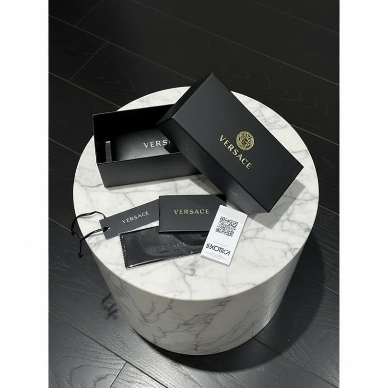 Puede incluir: Una caja negra de Versace con letras y logotipo dorados, abierta para revelar gafas de sol. Tambi&eacute;n hay etiquetas adicionales y un pa&ntilde;o de limpieza con el logotipo de Versace. Los art&iacute;culos se exhiben sobre una mesa de m&aacute;rmol blanco.