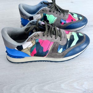 May include: A pair of colorful sneakers with a camouflage pattern. The shoes feature a mix of pink, green, blue, and brown hues. The soles are white and the laces are olive green.