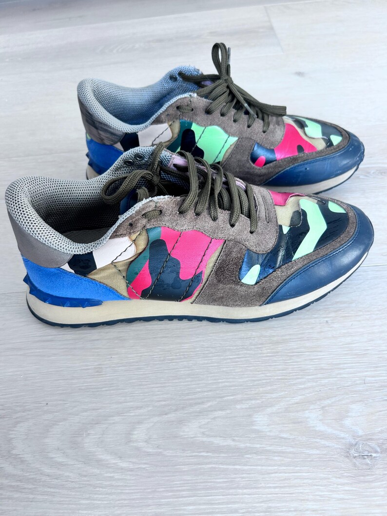 May include: A pair of colorful sneakers with a camouflage pattern. The shoes feature a mix of pink, green, blue, and brown hues. The soles are white and the laces are olive green.