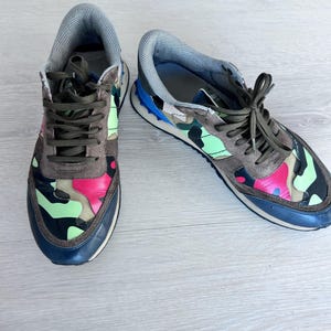 May include: A pair of sneakers with a camouflage pattern in shades of brown, pink, green, and black. The sneakers have brown laces, a gray tongue, and a navy blue sole. The shoes are on a light wood surface.