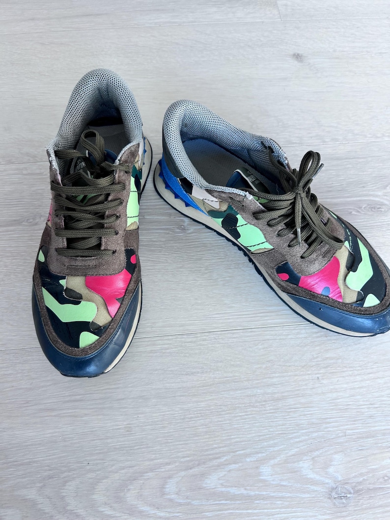 May include: A pair of sneakers with a camouflage pattern in shades of brown, pink, green, and black. The sneakers have brown laces, a gray tongue, and a navy blue sole. The shoes are on a light wood surface.