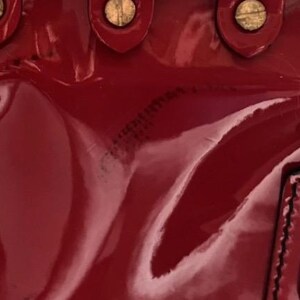 May include: Close-up of a glossy, deep red handbag. The bag features gold-toned hardware, including rivets and a clasp. The leather has a patent finish, reflecting light and creating highlights.