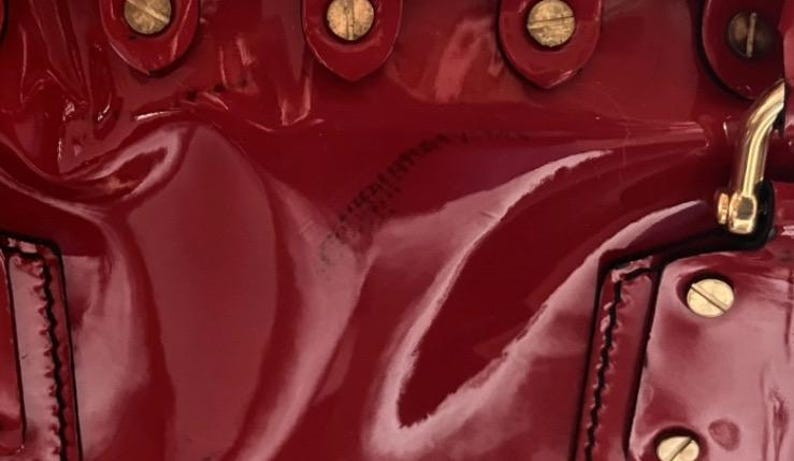 May include: Close-up of a glossy, deep red handbag. The bag features gold-toned hardware, including rivets and a clasp. The leather has a patent finish, reflecting light and creating highlights.