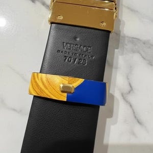 May include: Black leather belt with a gold-tone buckle and gold and blue accent. The belt is marked "VERSACE MADE IN ITALY 70/28". The belt's width is approximately 71 cm.