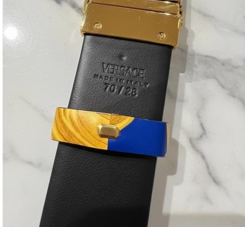 May include: Black leather belt with a gold-tone buckle and gold and blue accent. The belt is marked "VERSACE MADE IN ITALY 70/28". The belt's width is approximately 71 cm.