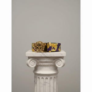 May include: A purple and gold patterned belt with a gold-coloured Medusa head buckle. The belt is displayed on a white marble column, showcasing its design and craftsmanship. The belt's pattern features a floral design.