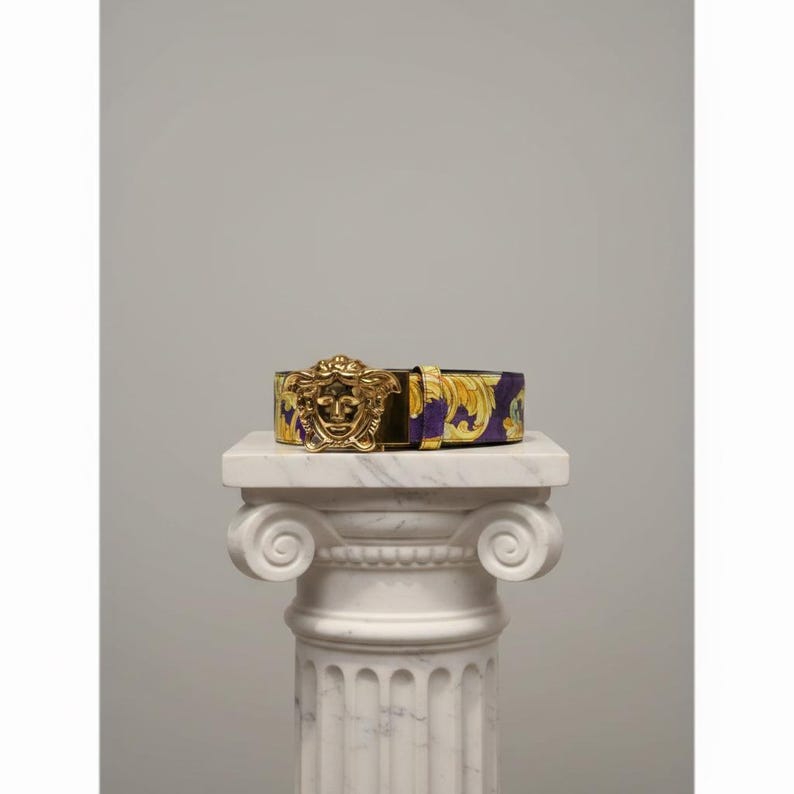 May include: A purple and gold patterned belt with a gold-coloured Medusa head buckle. The belt is displayed on a white marble column, showcasing its design and craftsmanship. The belt's pattern features a floral design.