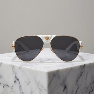 Puede incluir: Gafas de sol de aviador blancas y doradas con lentes oscuras. Las gafas tienen un puente y patillas blancos, con detalles dorados y un emblema dorado en el puente. Las gafas se exhiben sobre una superficie de m&aacute;rmol blanco.