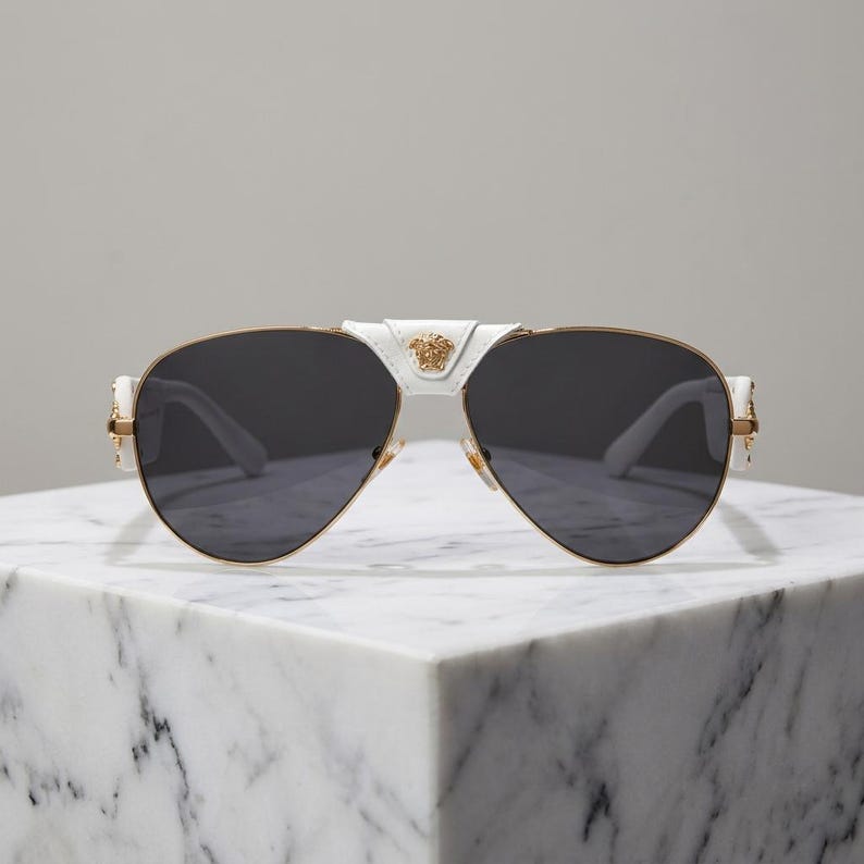 Puede incluir: Gafas de sol de aviador blancas y doradas con lentes oscuras. Las gafas tienen un puente y patillas blancos, con detalles dorados y un emblema dorado en el puente. Las gafas se exhiben sobre una superficie de m&aacute;rmol blanco.