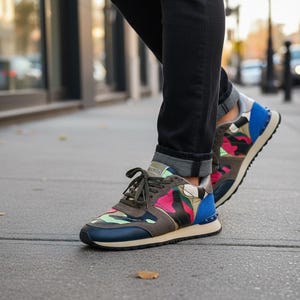 May include: Camouflage-patterned sneakers with blue, pink, and green accents. The sneakers have a navy blue toe cap, a beige sole, and blue detailing on the heel. The laces are olive green. The shoes are worn with black jeans.