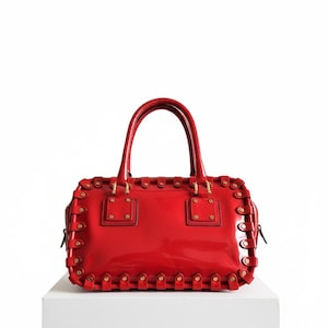 May include: A glossy red handbag with a rectangular shape and two top handles. The bag features decorative stitching and gold-colored hardware. It is displayed on a white surface against a white background.