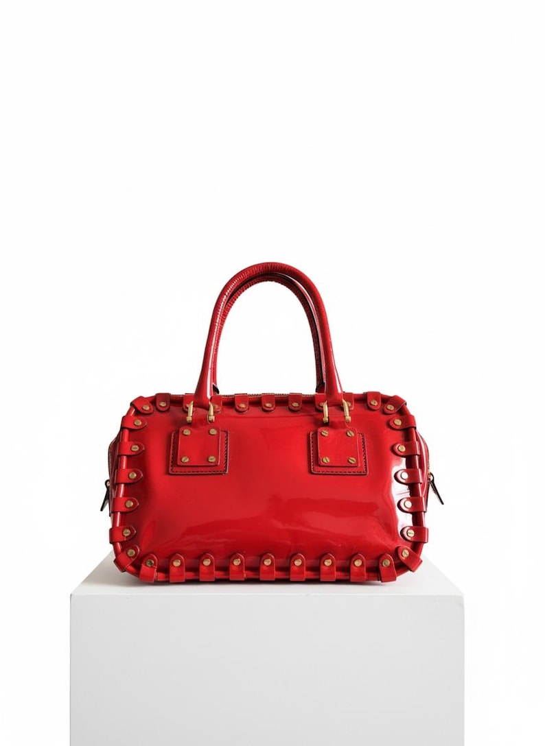 May include: A glossy red handbag with a rectangular shape and two top handles. The bag features decorative stitching and gold-colored hardware. It is displayed on a white surface against a white background.