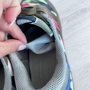 May include: Close-up of a multi-colored sneaker. The shoe features a camouflage pattern in shades of brown, green, and pink. The interior is light gray, and the laces are olive green. The sole has blue and gray accents.